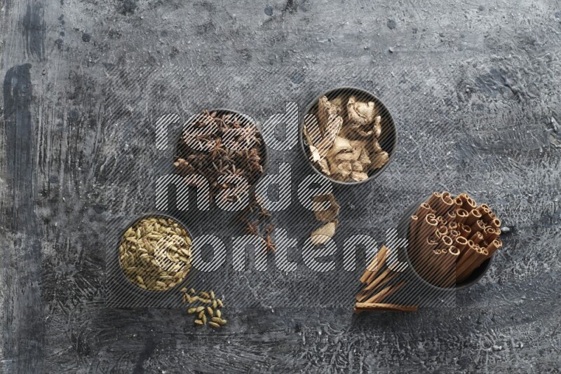 Ginger, Cardamom, Star anise and cinnamon sticks in 4 bowls on a textured black background