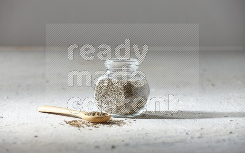 A glass spice jar and wooden spoon full of cumin seeds on textured white flooring