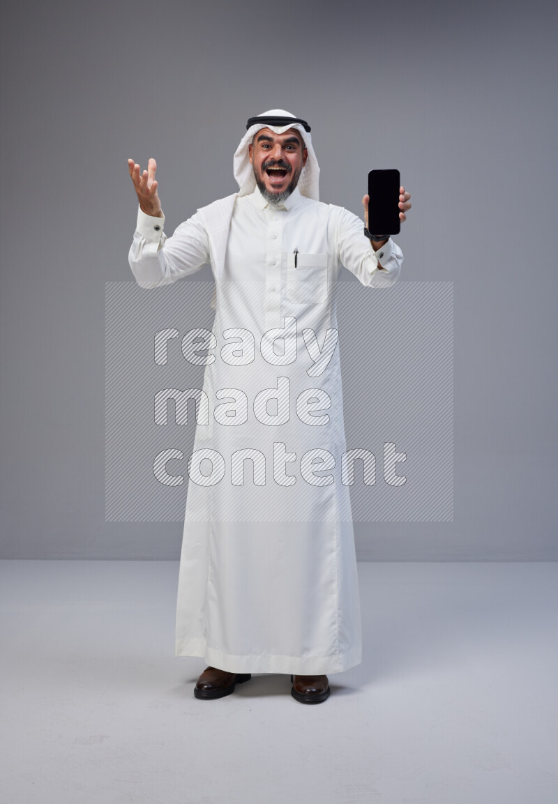 Saudi man Wearing Thob and white Shomag standing showing phone to camera on Gray background