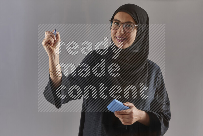 Woman in black abaya, hijab and glasses holding a marker pen to write on a transparent board with different reactions and expressions on grey background