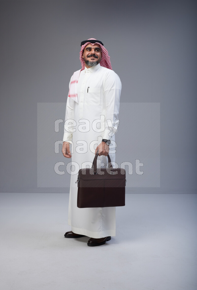 Saudi man Wearing Thob and red Shomag standing holding bag on Gray background