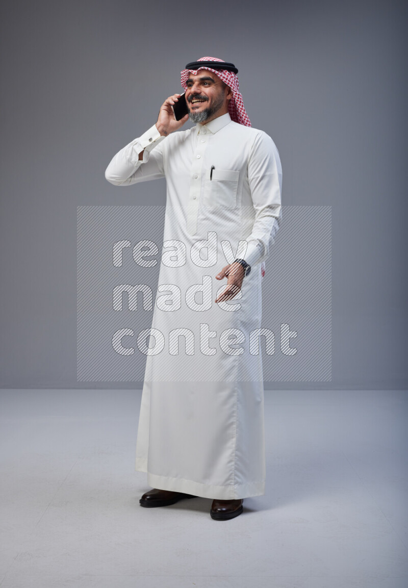 Saudi man Wearing Thob and red Shomag standing talking on phone on Gray background
