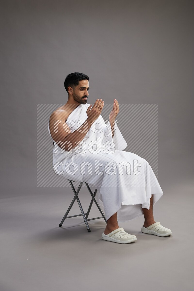 A man wearing Ehram sitting on chair performing dua'a on gray background