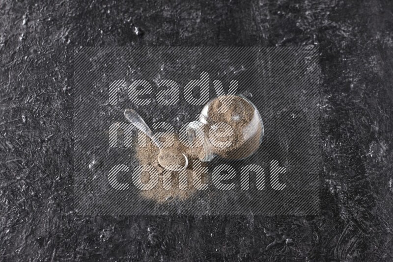 A flipped glass spice jar full of black pepper powder with a metal spoon full of powder on textured black flooring