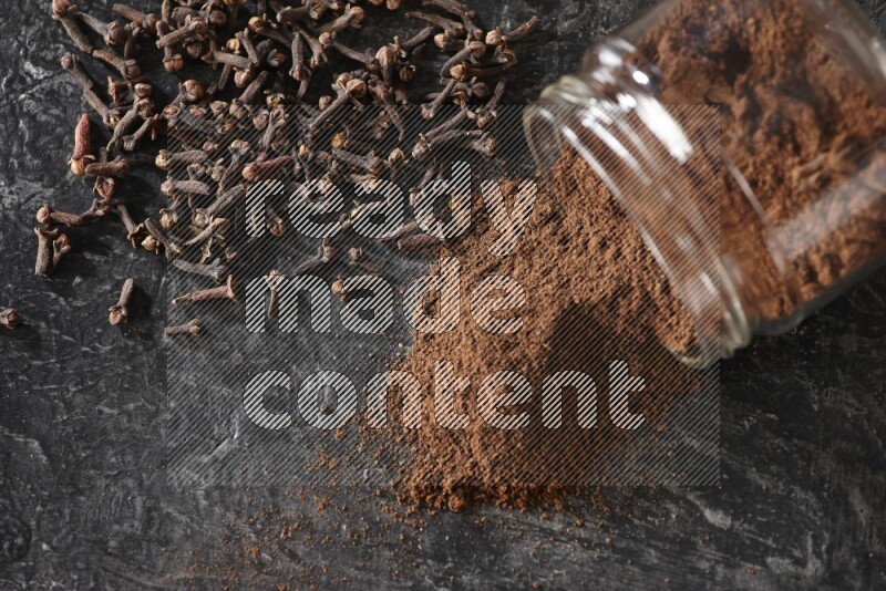 A flipped glass jar full of cloves powder with cloves spread on a textured black flooring