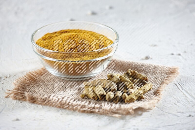 A glass bowl full of turmeric powder and dried turmeric whole finger on a piece of burlap on a textured white flooring