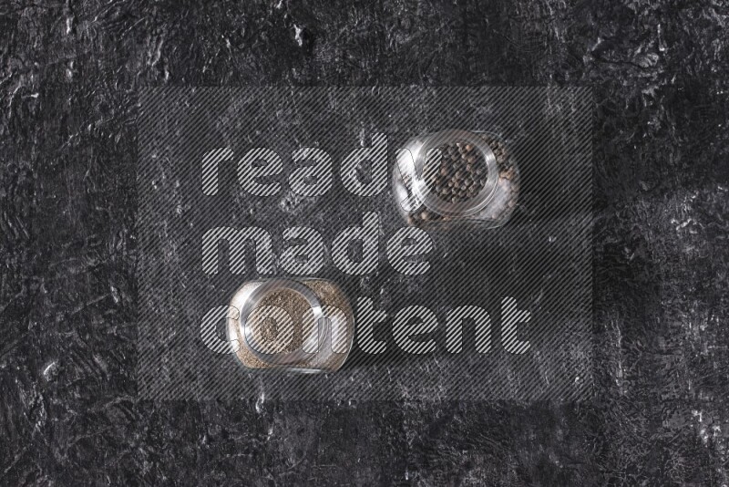 2 glass spice jars full of black pepper powder and black pepper beads on textured black flooring