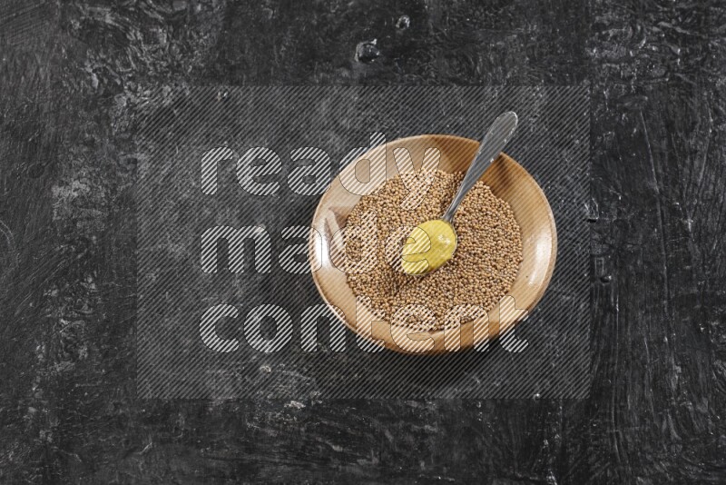 A beige pottery plate filled with mustard seeds with a metal spoon full of mustard paste in it on a textured black flooring