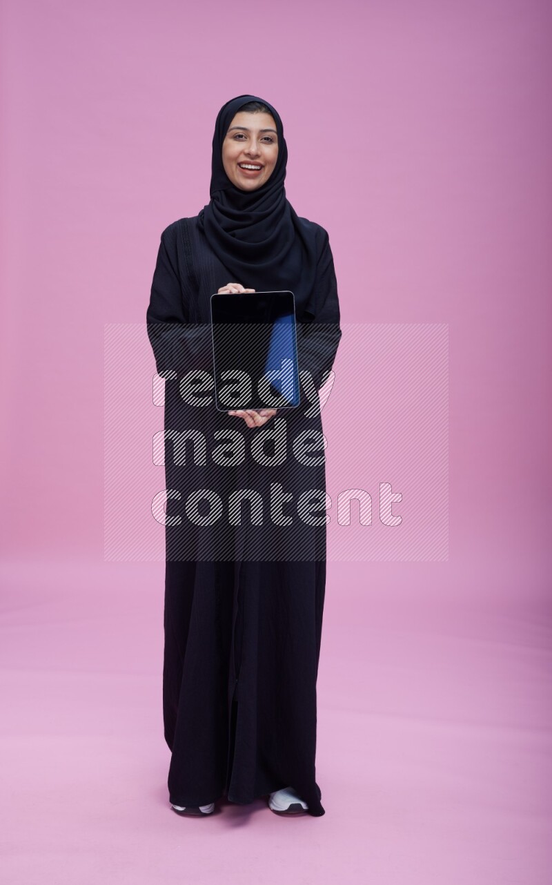 Saudi woman wearing Abaya standing showing tablet to camera on pink background