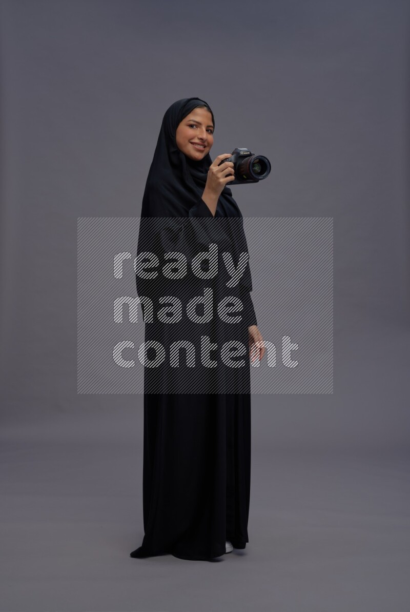 Saudi woman wearing Abaya standing holding Camera on gray background