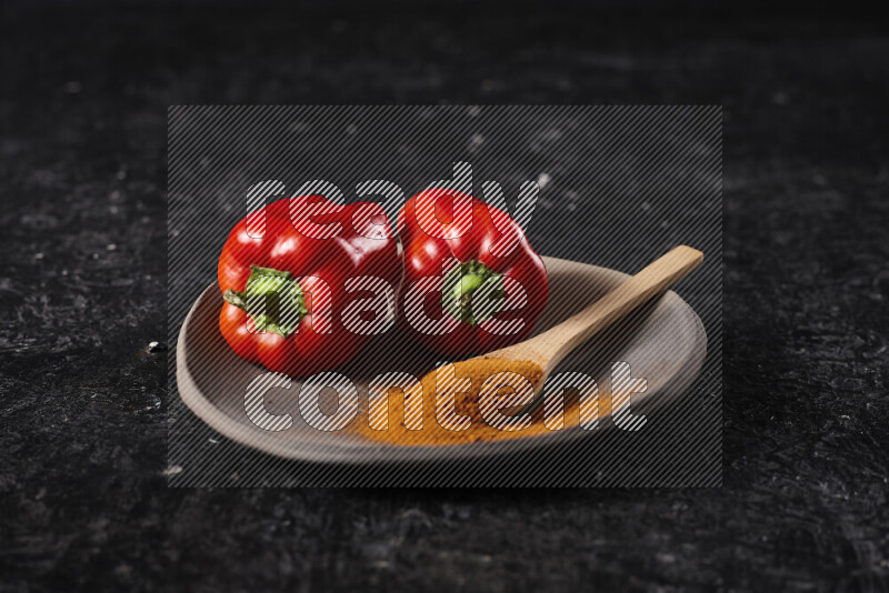 A wooden spoon full of ground paprika powder with red bell peppers, all on a pottery plate on black background