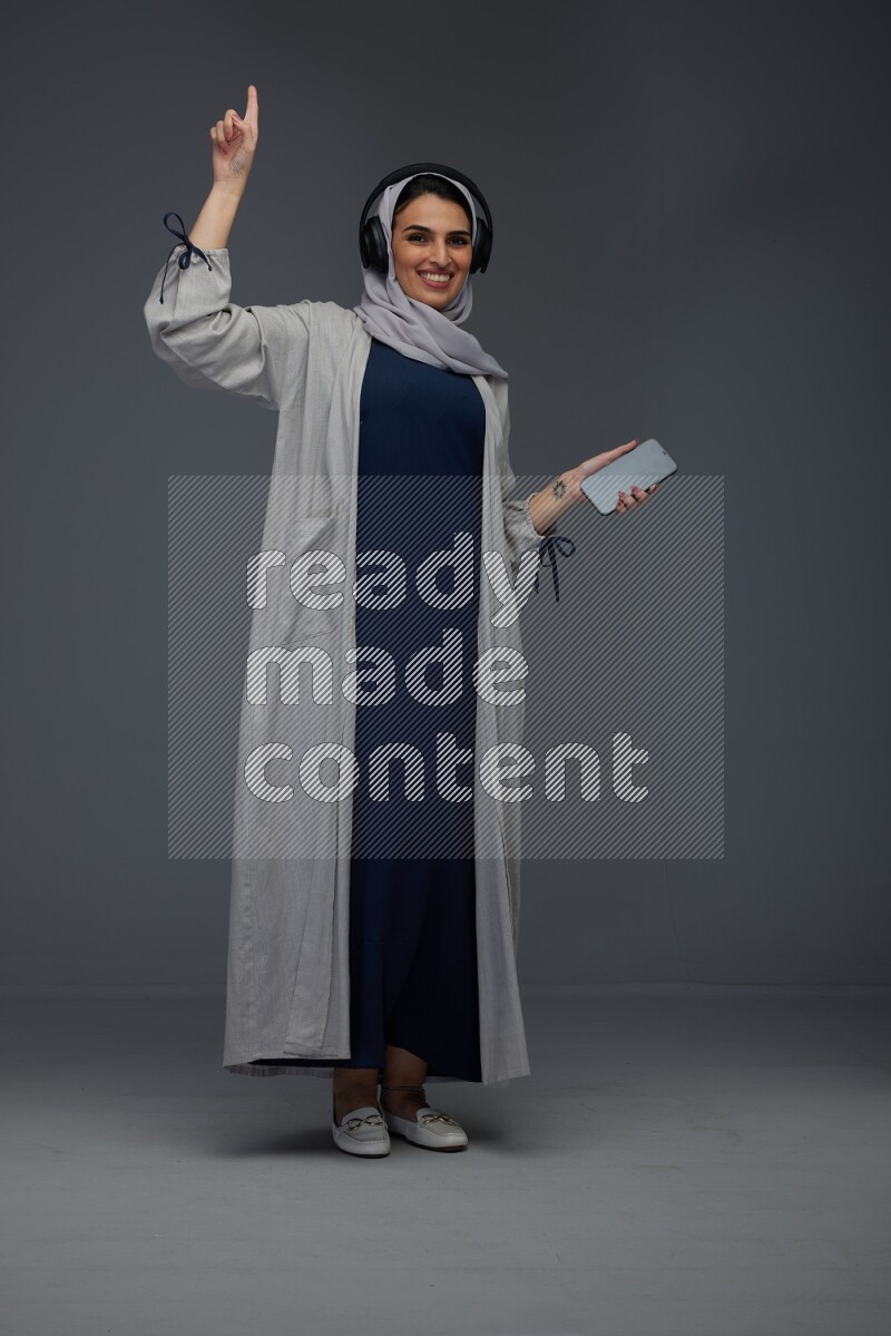 A Saudi woman wearing a light gray Abaya and head scarf standing and listening to music on a grey background