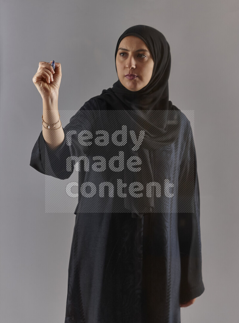 Woman in black abaya and hijab holding a marker pen to write on a transparent board with different reactions and expressions on grey background