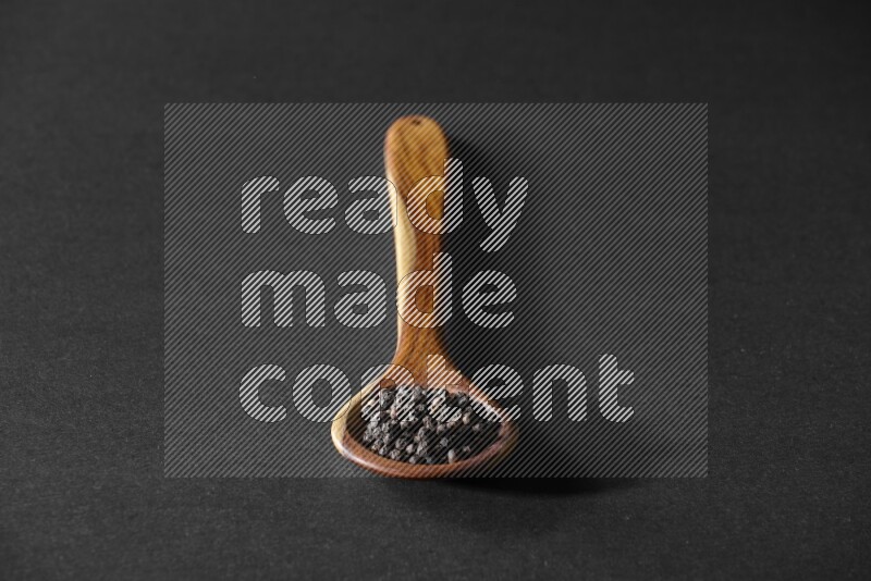 A wooden ladle full of black pepper beads on a black flooring