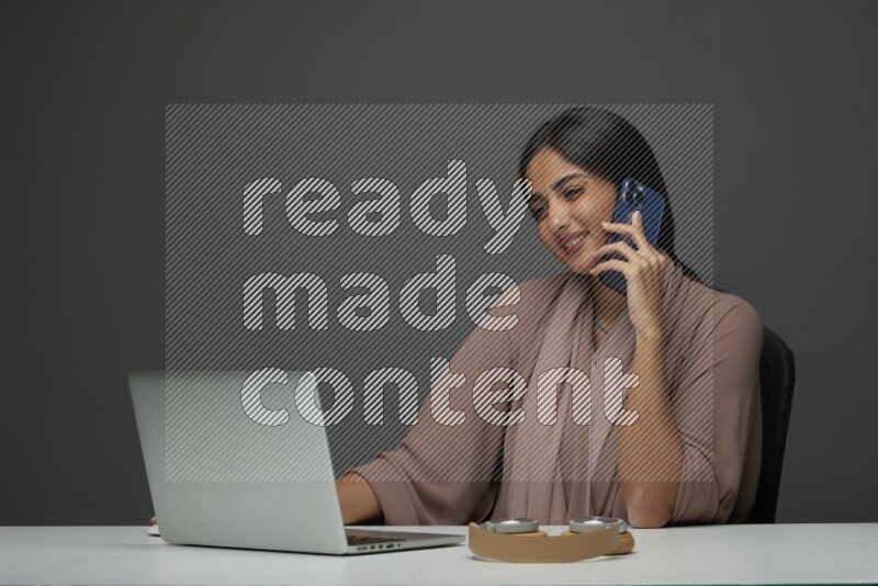 A Saudi woman Sitting on her desk Calling  on a Gray Background wearing Brown Abaya