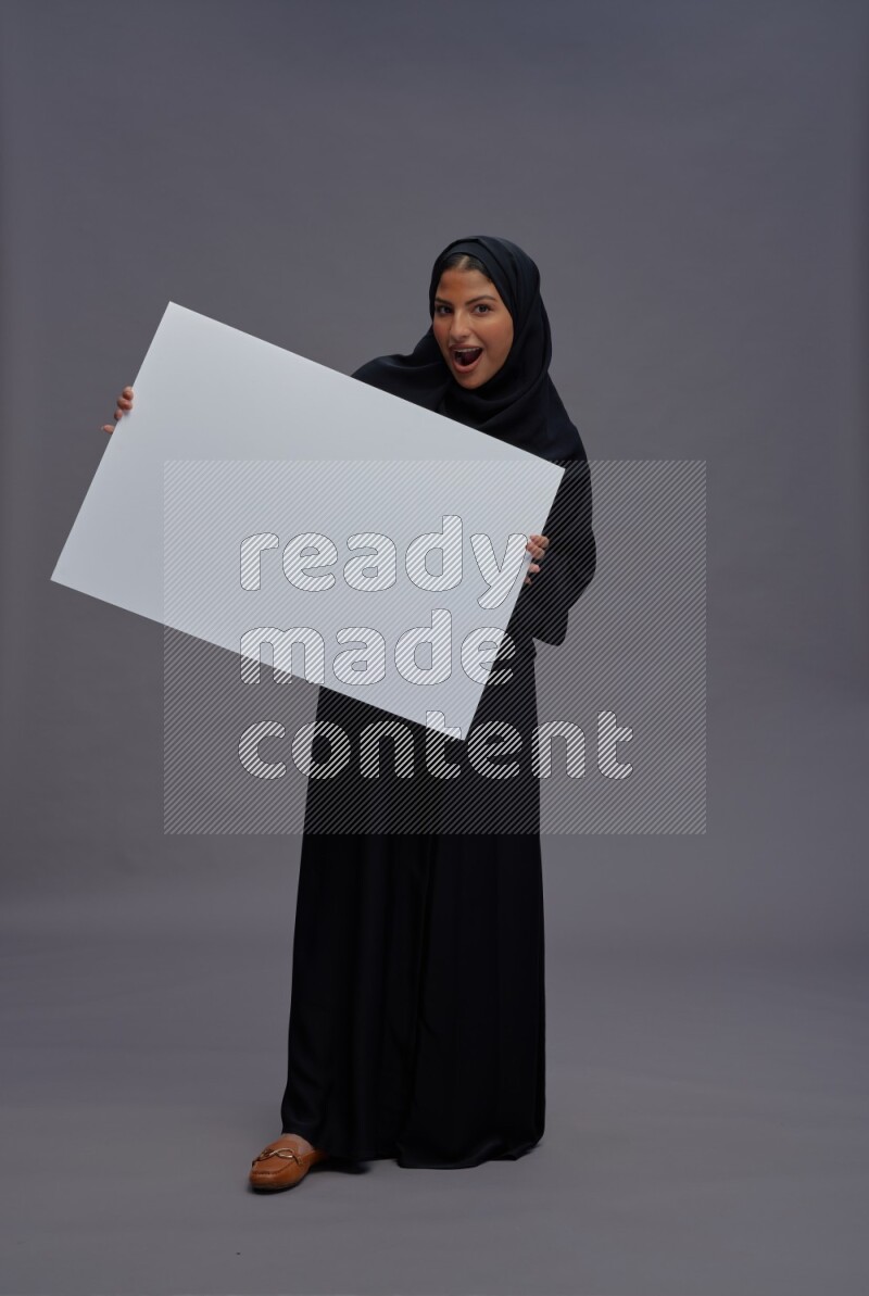 Saudi woman wearing Abaya standing holding white board on gray background