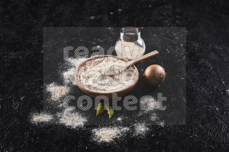A wooden bowl full of onion powder with a glass jar beside it and fresh onion on black background
