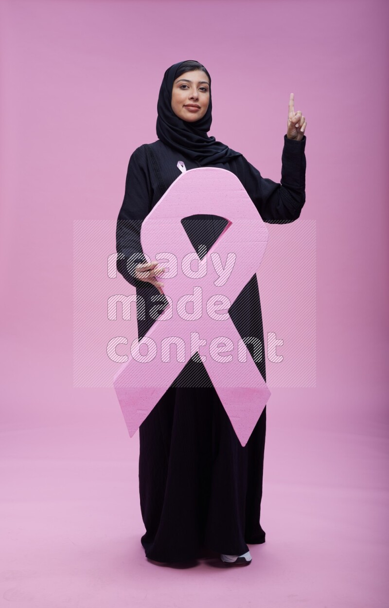 Saudi woman wearing pink ribbon on abaya standing holding awareness ribbon on plain pink background