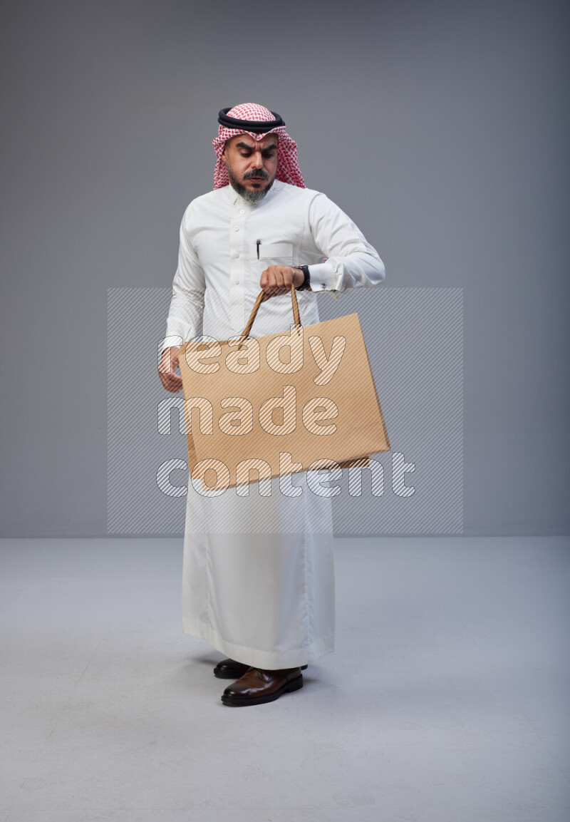 Saudi man Wearing Thob and red Shomag standing holding shopping bag on Gray background