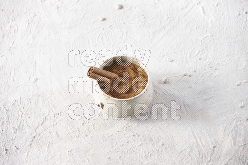Ceramic beige bowl full of cinnamon powder with a cinnamon stick on a textured white background