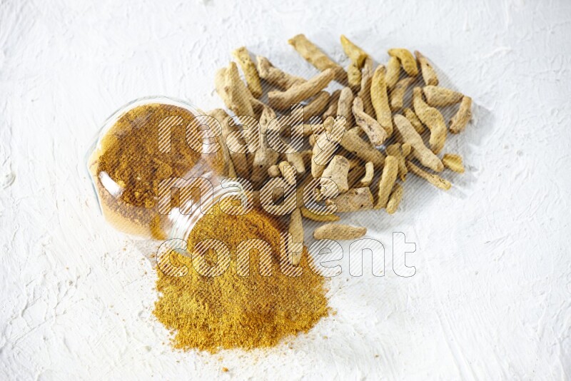 A flipped glass spice jar full of turmeric powder and powder spilled out of it with dried whole fingers on textured white flooring