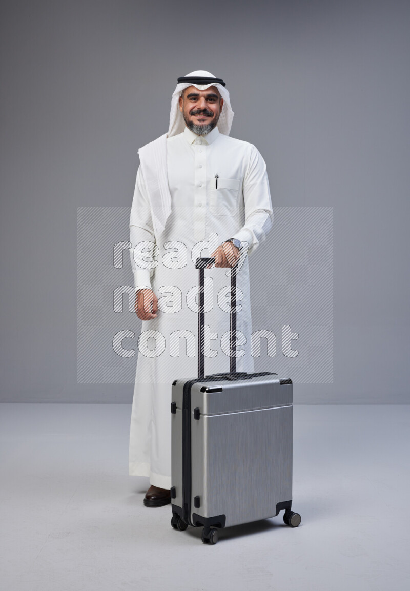 Saudi man wearing Thob and white Shomag standing holding Travel bag on Gray background
