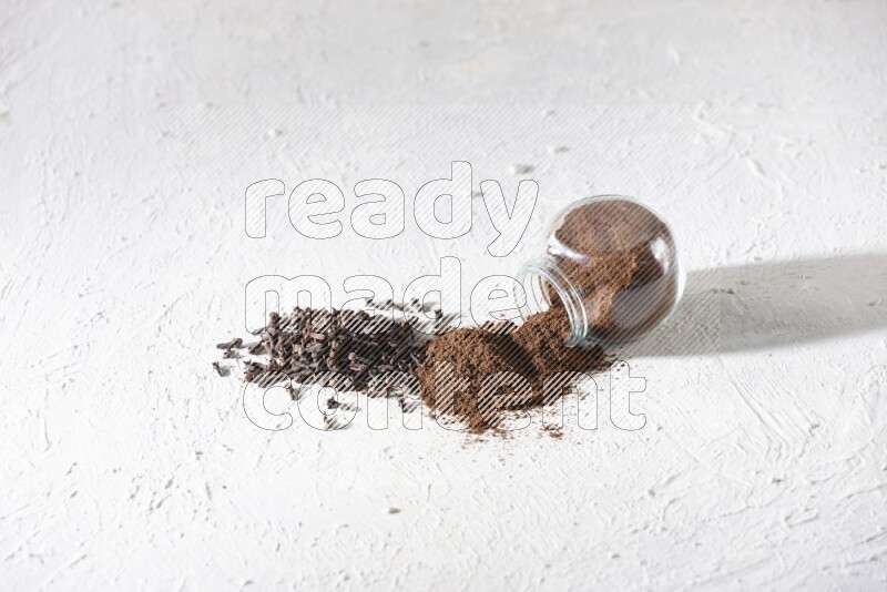 وعاء زجاجي للتوابل مقلوب ممتلئ ببودرة القرنفل وتناثرت البودرة منه مع حبوب القرنفل المنتشرة على أرضية بيضاء