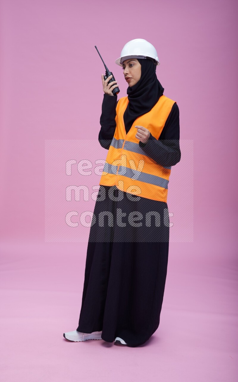 Saudi woman wearing Abaya with engineer vest and helmet standing holding walkie-talkie on pink background