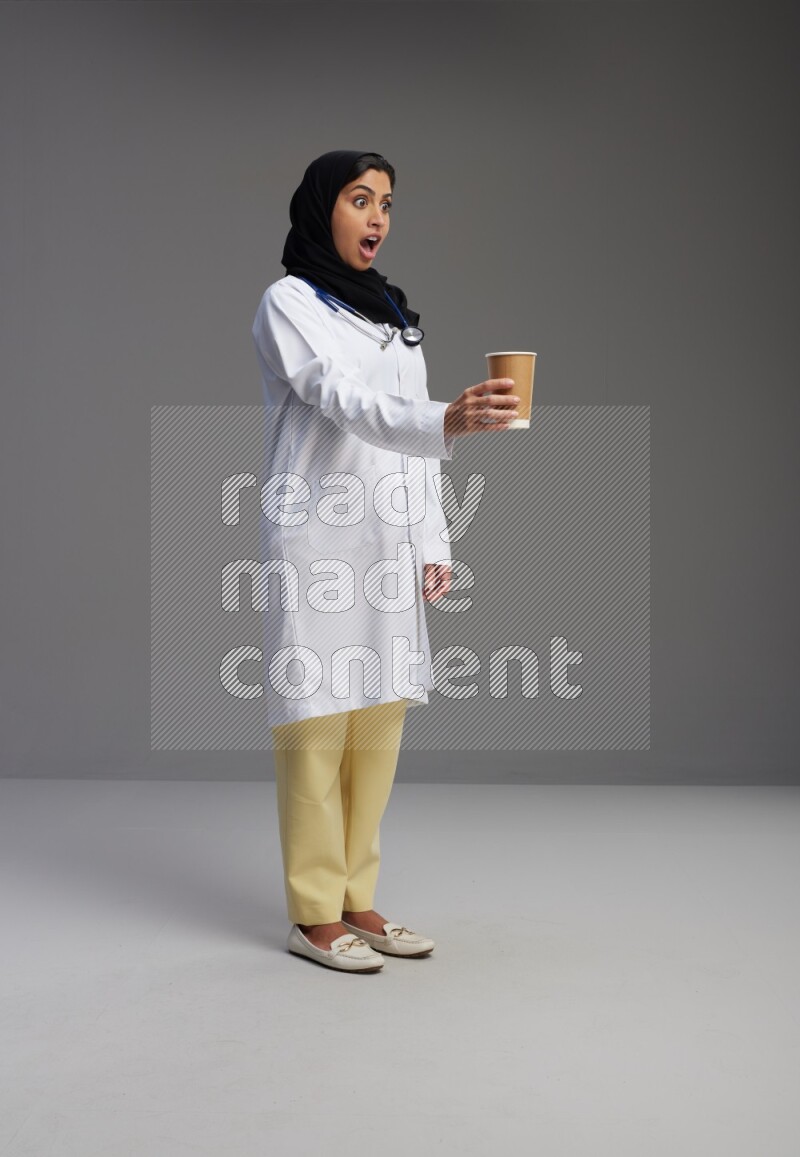 Saudi woman wearing lab coat with stethoscope standing holding paper cup on Gray background