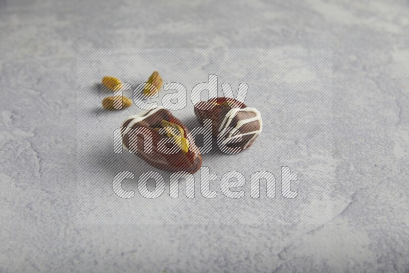 two pistachio stuffed dates covered with dark chocolate with white chocolate drizzle with unroasted pistachios on a light grey background