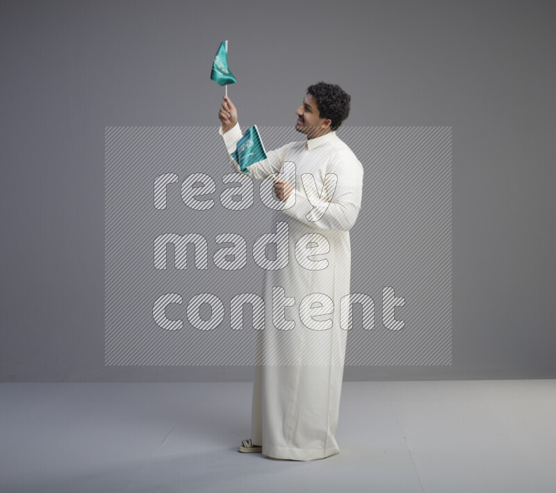 A Saudi man standing wearing thob holding small Saudi flag on gray background