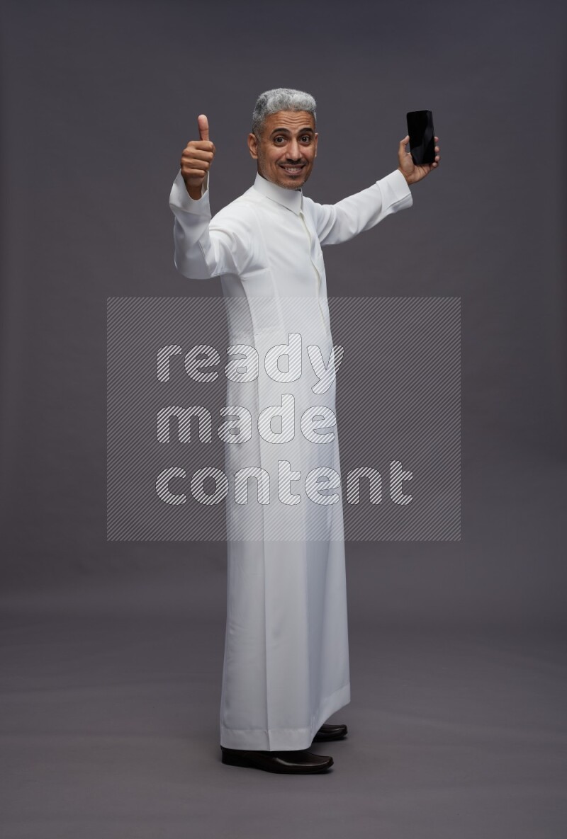 Saudi man wearing thob standing taking selfie on gray background
