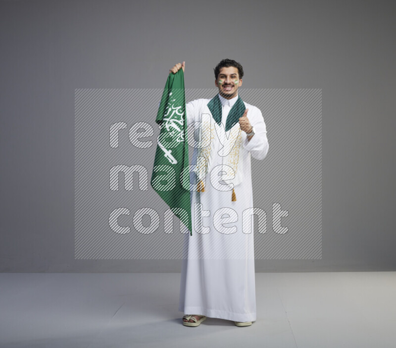 A Saudi man standing wearing thob with face painting wrapping Saudi flag scarf and holding big Saudi flag on gray background