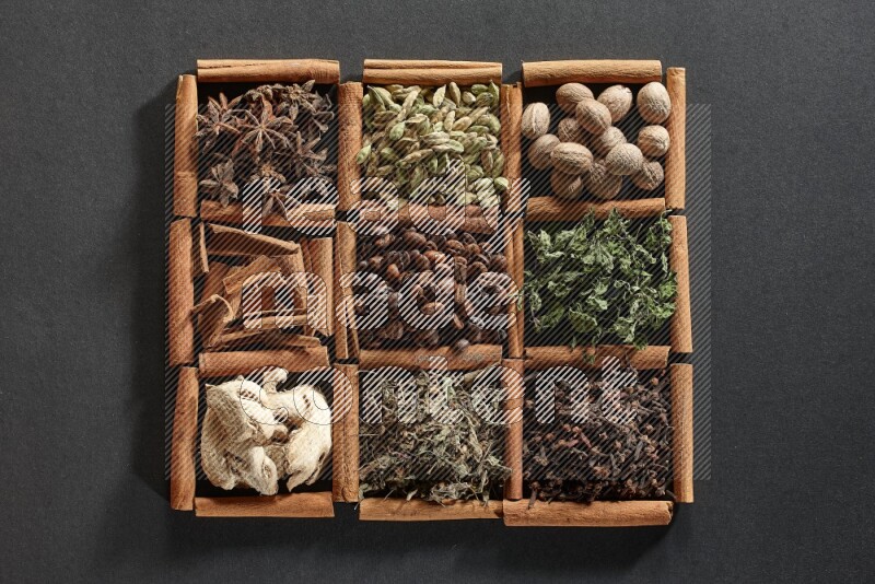 9 squares of cinnamon sticks full of coffee beans in the middle surrounded by nutmeg, dried mint, cloves, dried basil, dried ginger, cinnamon, star anise and cardamom on black flooring
