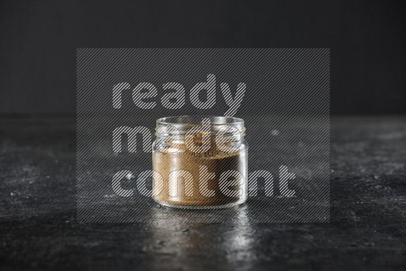 A glass jar full of cumin powder on a textured black flooring
