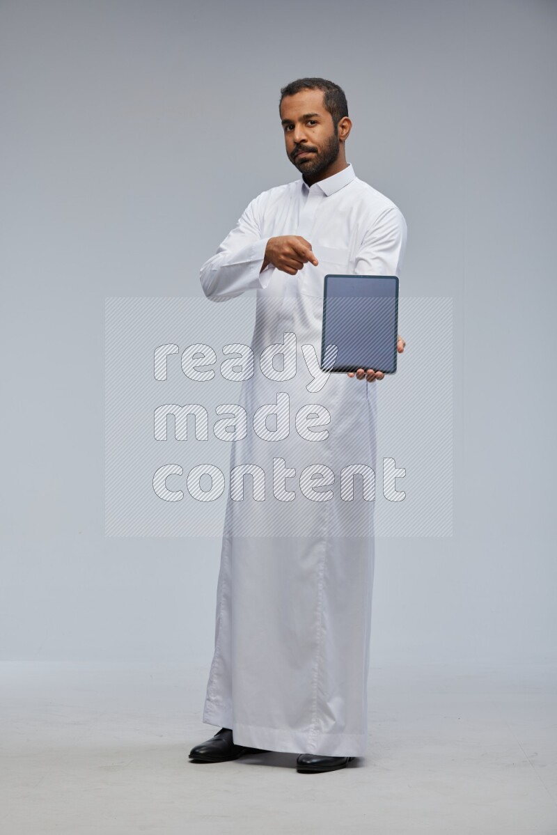 Saudi man wearing Thob standing showing tablet to camera on Gray background