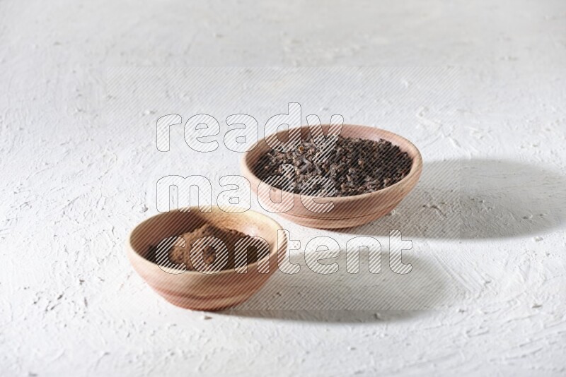2 wooden bowls full of cloves powder and whole cloves on a textured white flooring