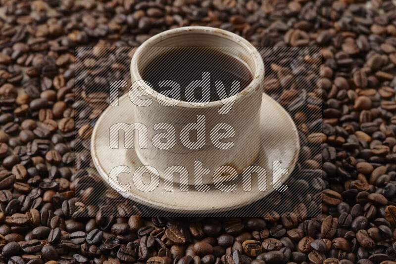A beige pottery cup of coffee surrounded by roasted coffee beans on beige background