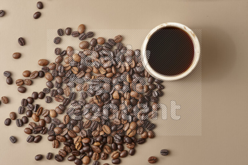 A beige pottery cup of coffee surrounded by roasted coffee beans on beige background