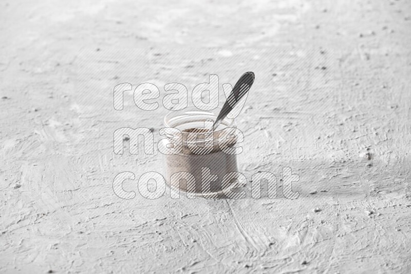 A glass jar full of black pepper powder and a metal spoon full of powder on a textured white flooring