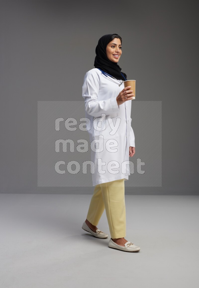 Saudi woman wearing lab coat with stethoscope standing holding paper cup on Gray background