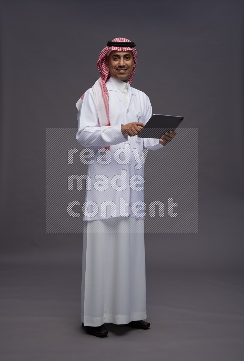 Saudi man wearing thob with lab coat and shomag with pocket employee badge standing working on tablet on gray background
