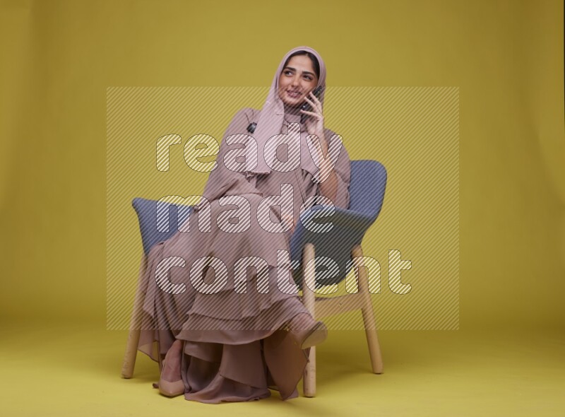 A woman having a call on a blue Chair on a Yellow Background wearing Brown Abaya with Hijab