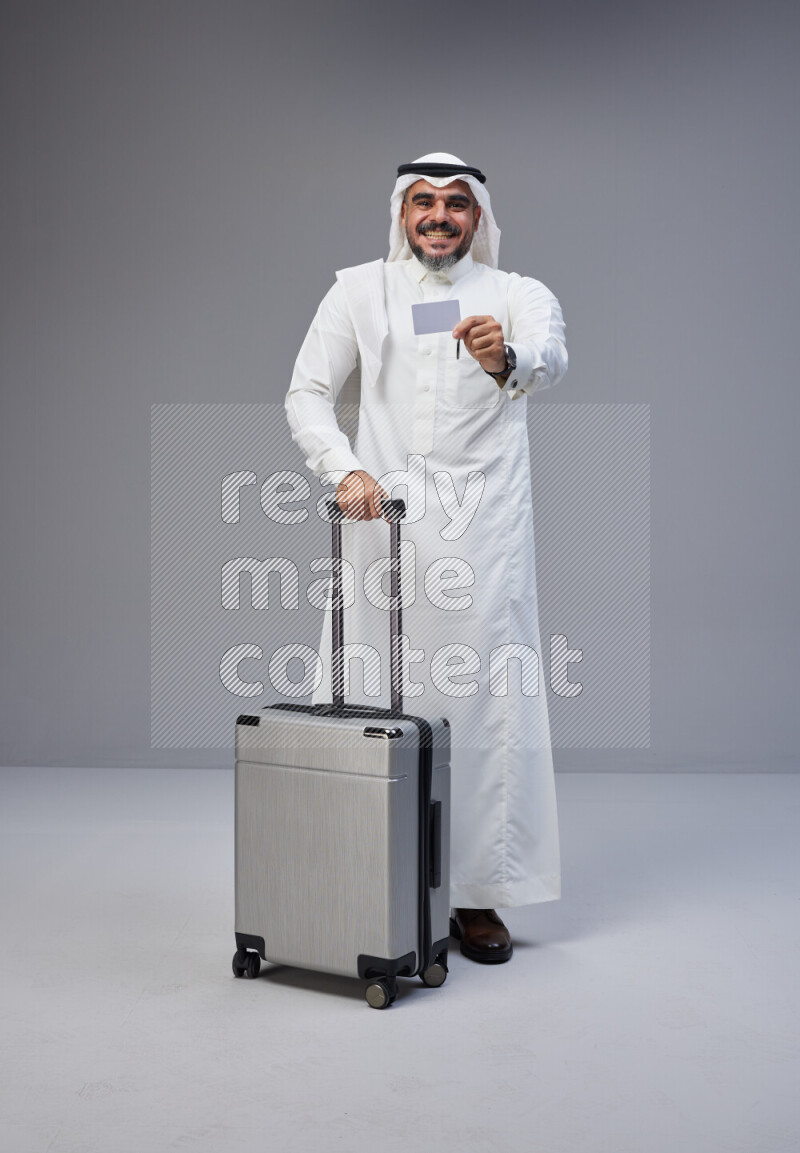 Saudi man wearing Thob and white Shomag standing holding Travel bag and ATM card on Gray background