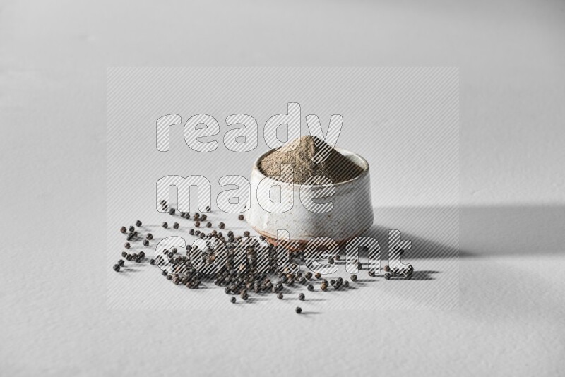A white ceramic bowl full of black pepper powder and black pepper beads spread on white flooring
