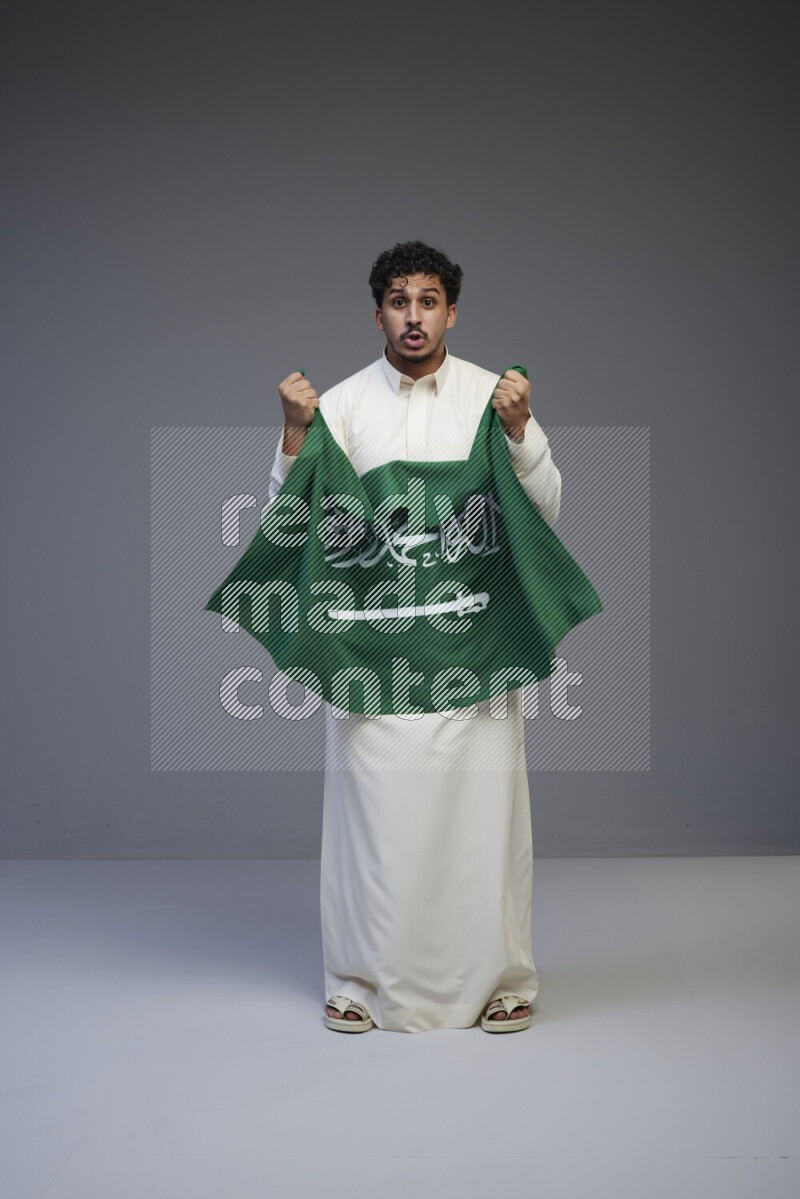 A Saudi man standing wearing thob holding big Saudi flag on gray background