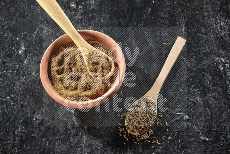 A wooden bowl and 2 wooden spoons full of cumin powder and cumin seeds on a textured black flooring