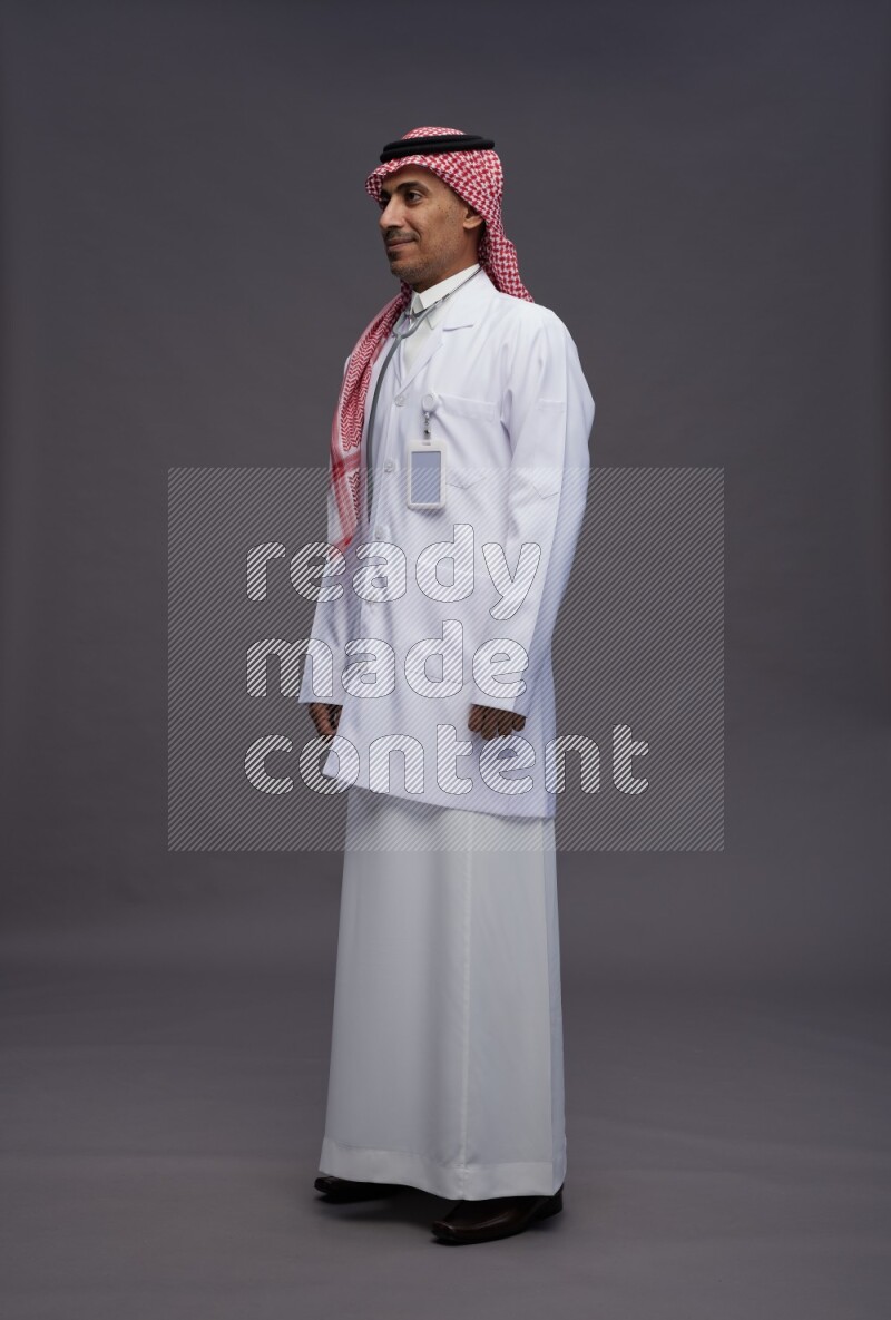 Saudi man wearing thob with lab coat and shomag with pocket employee badge with stethoscope standing interacting with the camera on gray background