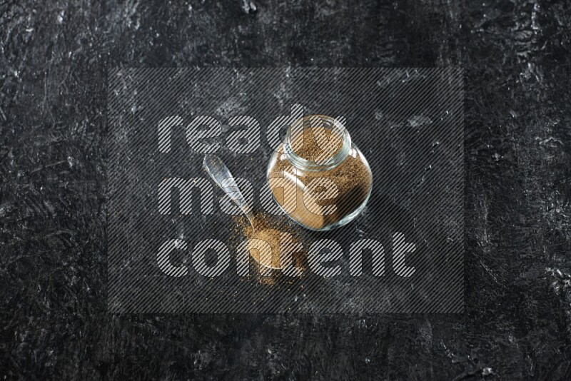 A glass spice jar and a metal spoon full of cumin powder on a textured black flooring