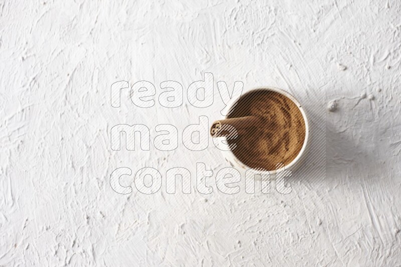 Ceramic beige bowl full of cinnamon powder with a cinnamon stick on a textured white background